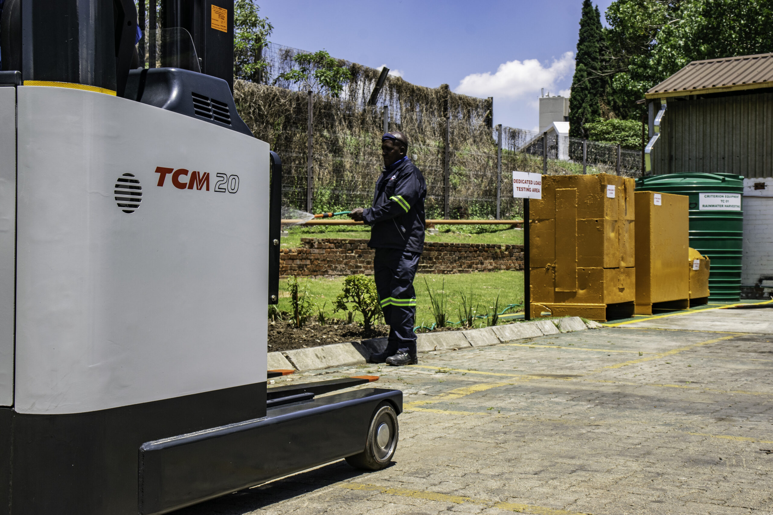 Operator standing near a TCM reach truck forklift at an outdoor worksite with containers and fencing in the background